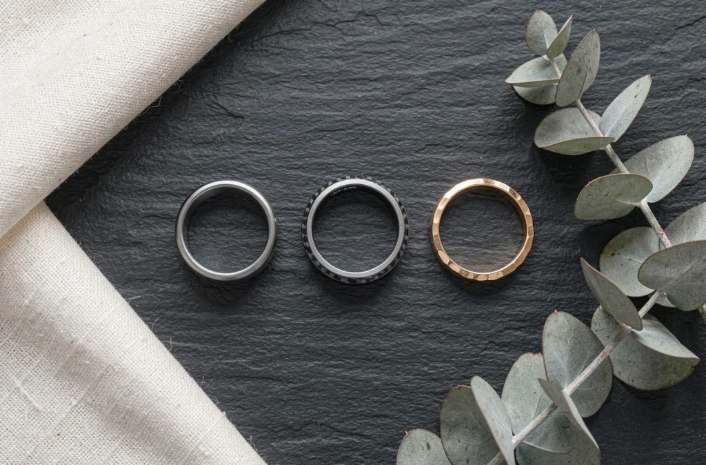 Flat lay de trois styles de bagues de PACS pour homme — tungstène, fibre de carbone et vermeil martelé — disposées sur une ardoise sombre avec lin et eucalyptus